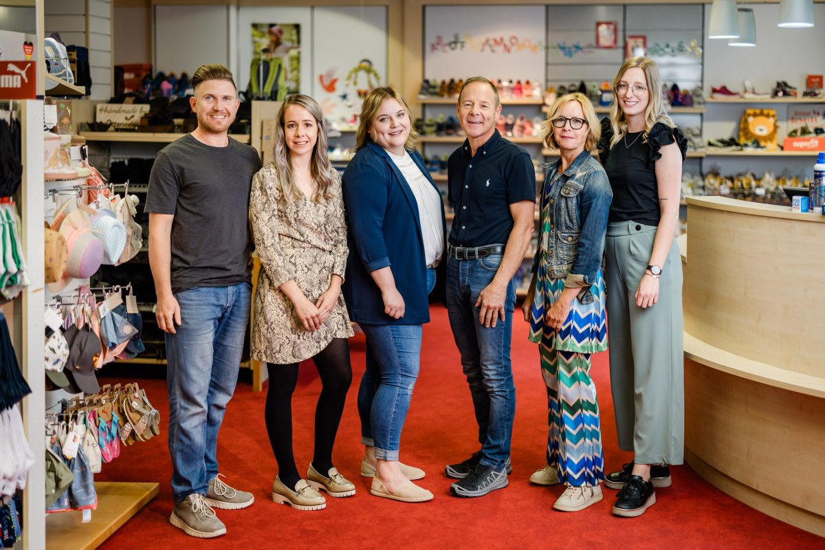 Gruppenfoto von den sechs Mitarbeiter:innen im Schuhgeschäft, sie stehen vor Regalen mit Schuhen und lächeln in die Kamera.