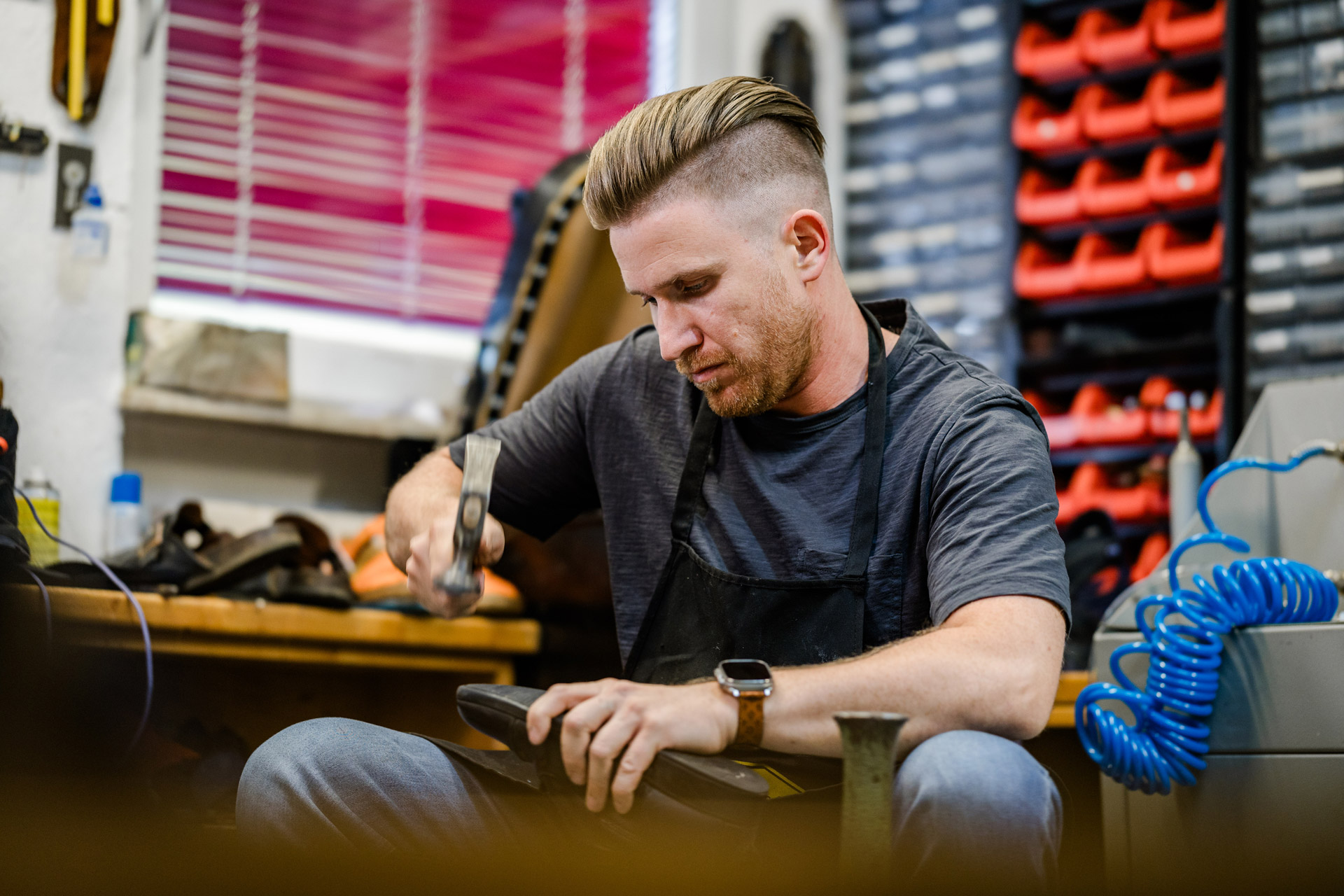 Schuhmacher in einer Werkstatt arbeitet mit einem Hammer an einem Schuh. Im Hintergrund stehen Regale mit Materialien und Werkzeug.