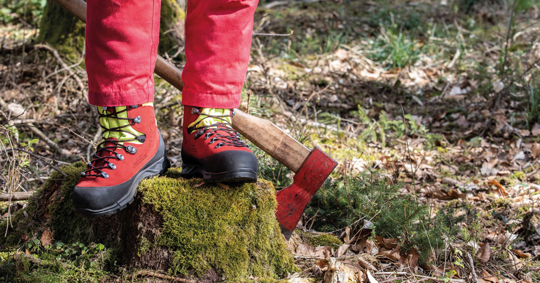 Person in roten Hosen steht mit rot-gelben Arbeitsschuhen auf einem moosigen Baumstumpf im Wald; daneben liegt eine Axt mit Holzstiel am Boden.