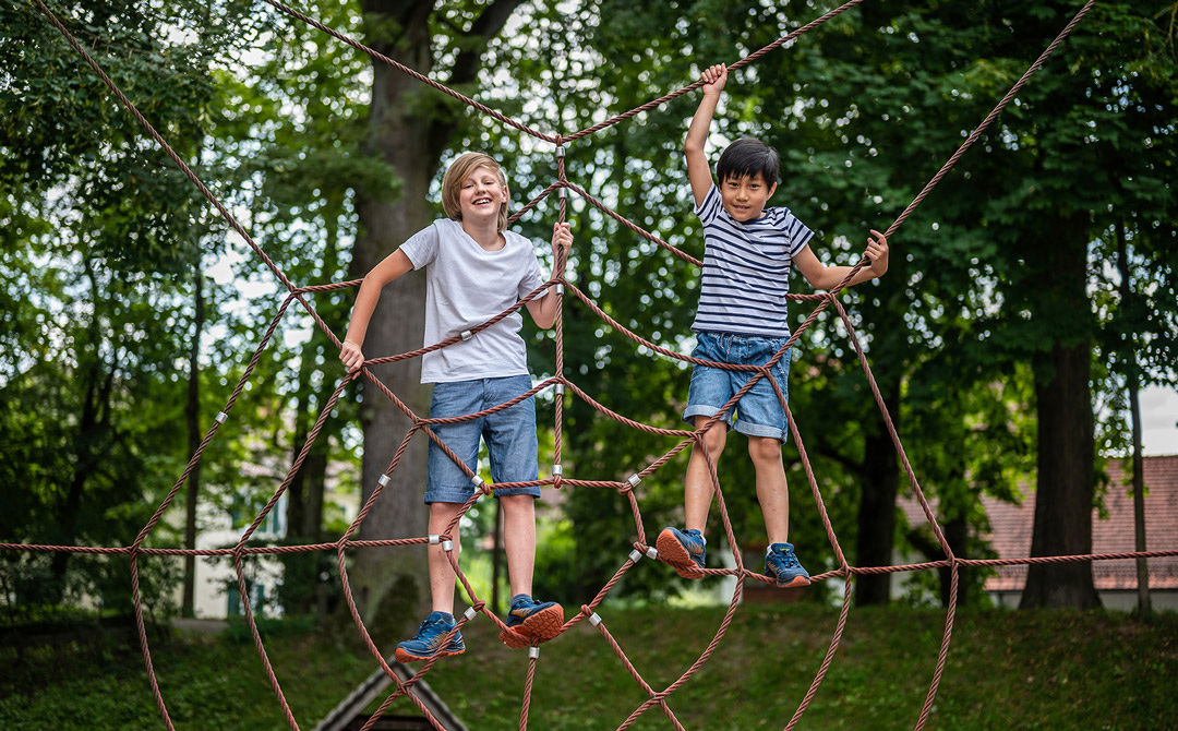 Zwei Kinder klettern und stehen in einem Seil-Kletternetz auf einem Spielplatz, im Hintergrund stehen große Bäume.