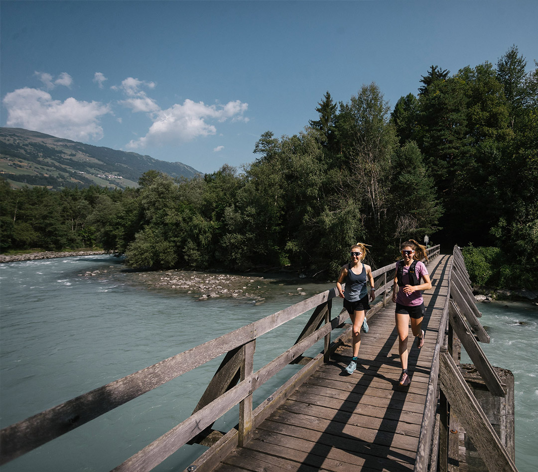 Zwei Frauen joggen über eine Holzbrücke neben einem Fluss, im Hintergrund sind Berge und ein Wald unter blauem Himmel.
