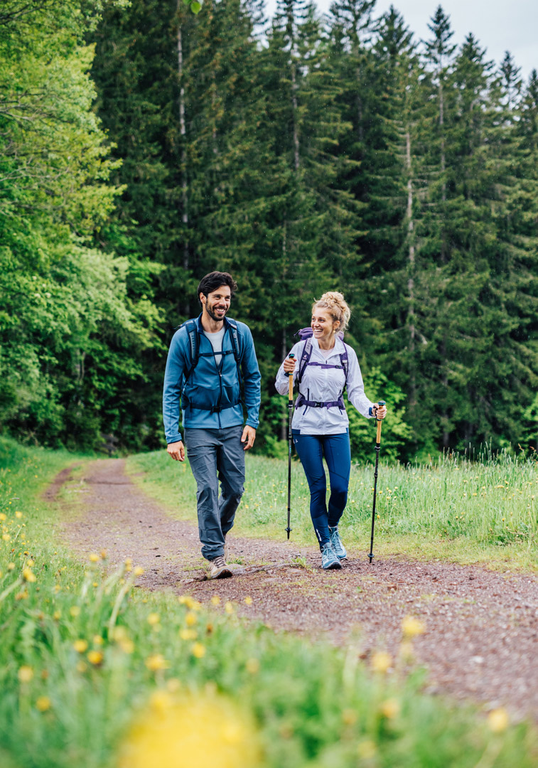 Wanderndes Paar mit Rucksäcken und Trekkingstöcken auf einem Weg, umgeben von dichtem Wald.