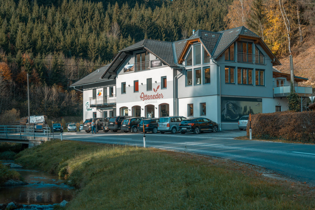 Schuhgeschäft Schuhhaus Atteneder. Zweistöckiges Haus, davor stehen Autos am Parkplatz. Im Hintergrund befindet sich ein Hang mit Bäumen und im Vordergrund ein Bach mit Wiese.