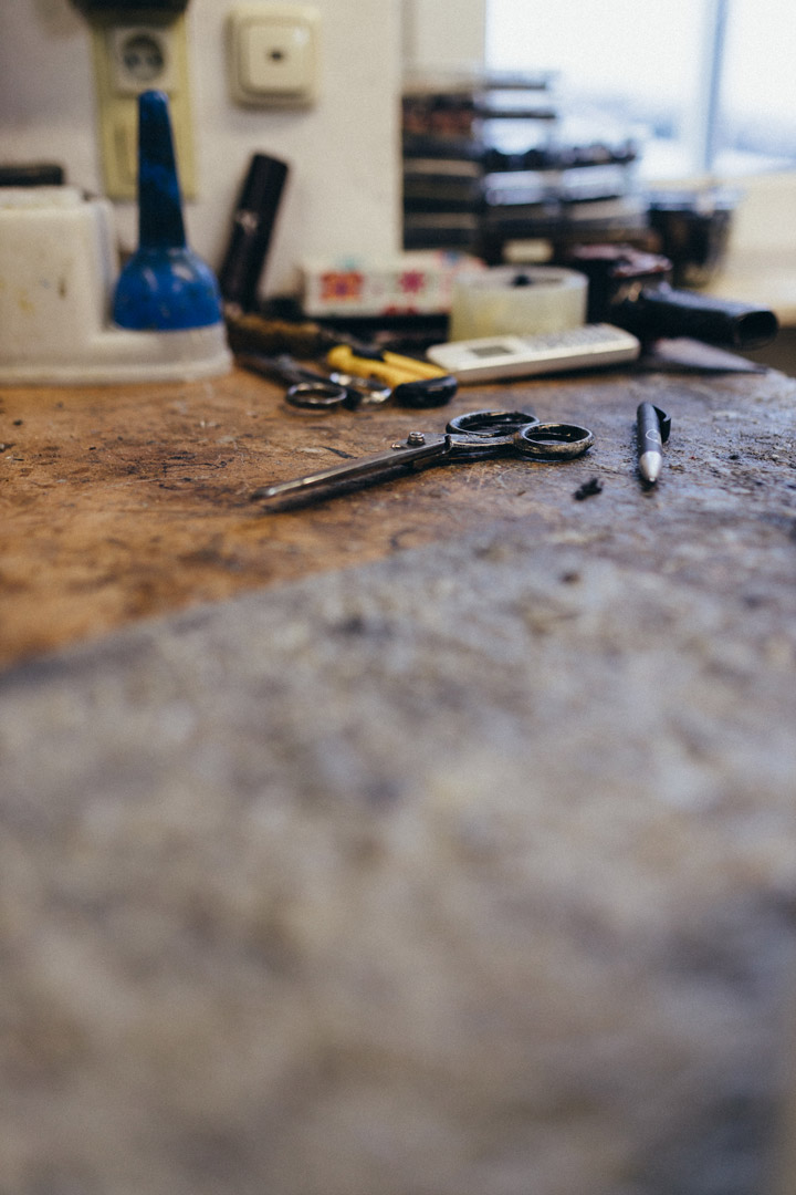 Schere und Stift liegen auf einer abgenutzten Werkbank. Im Hintergrund sind weitere Werkzeuge und Arbeitsmaterialien unscharf zu sehen.