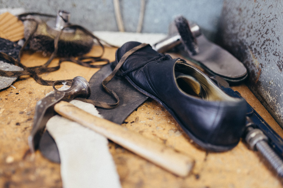 Nahaufnahme eines schwarzen Lederschuhs auf einer Werkbank, umgeben von Schuhmacherwerkzeug, Lederresten und Schnürsenkeln.